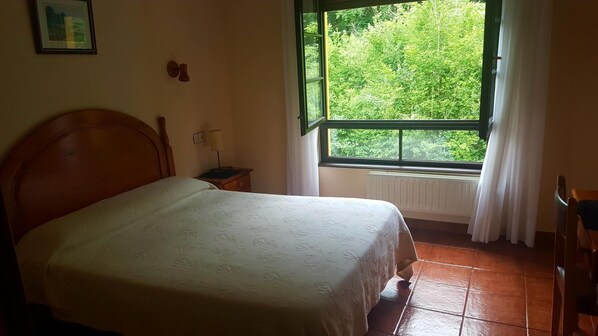 A bedroom with a wooden bed, a nightstand, a window with a view of greenery, and a radiator.