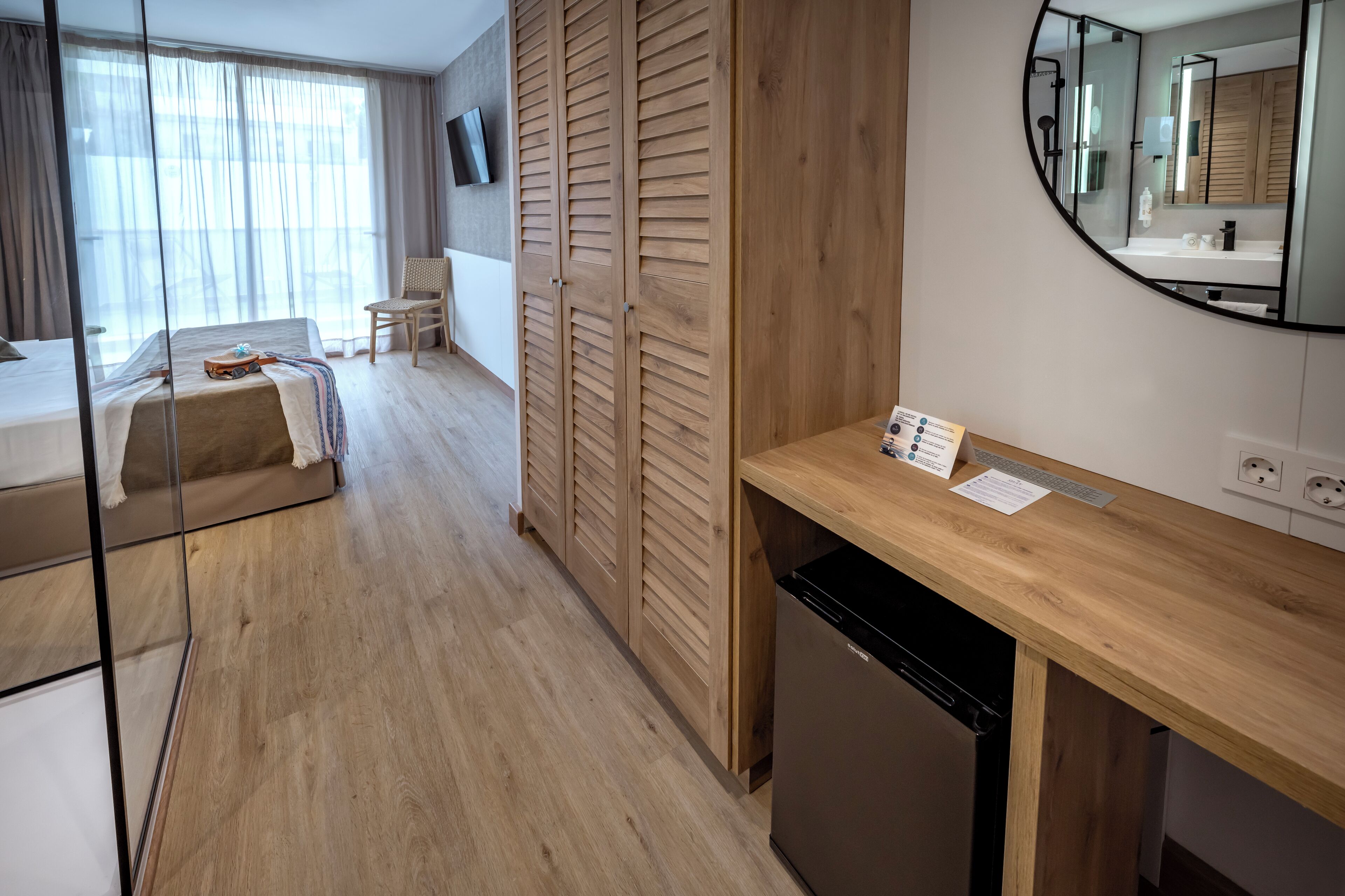 Quarto de hotel moderno com piso de madeira, uma cama com cobertura bege, guarda-roupa de madeira, pequena escrivaninha de madeira com espelho e televisão fixada na parede.