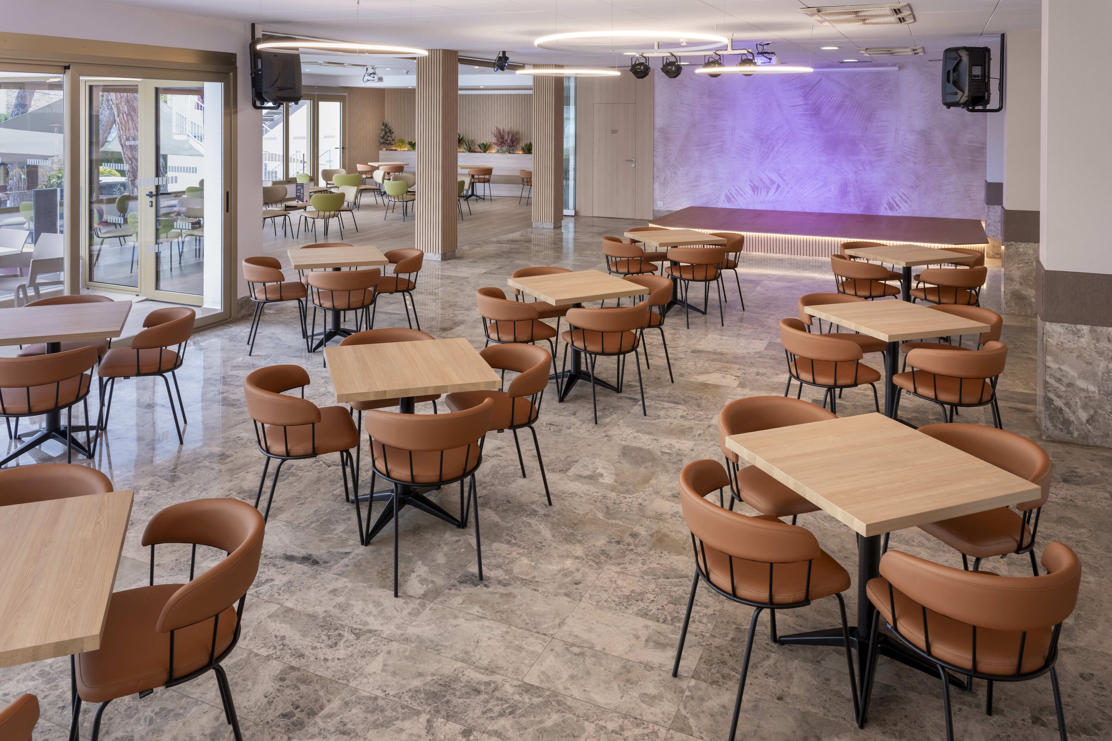 A spacious indoor dining area with multiple tables and chairs, a stage with a purple backdrop, and large windows allowing natural light.