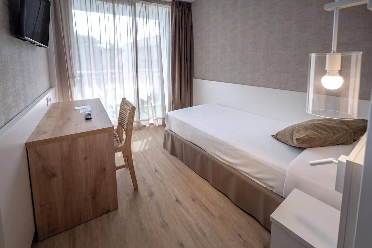 Een hotelkamer met een houten bureau, een enkel bed, een stoel, een aan de muur bevestigde televisie en een raam met gordijnen.