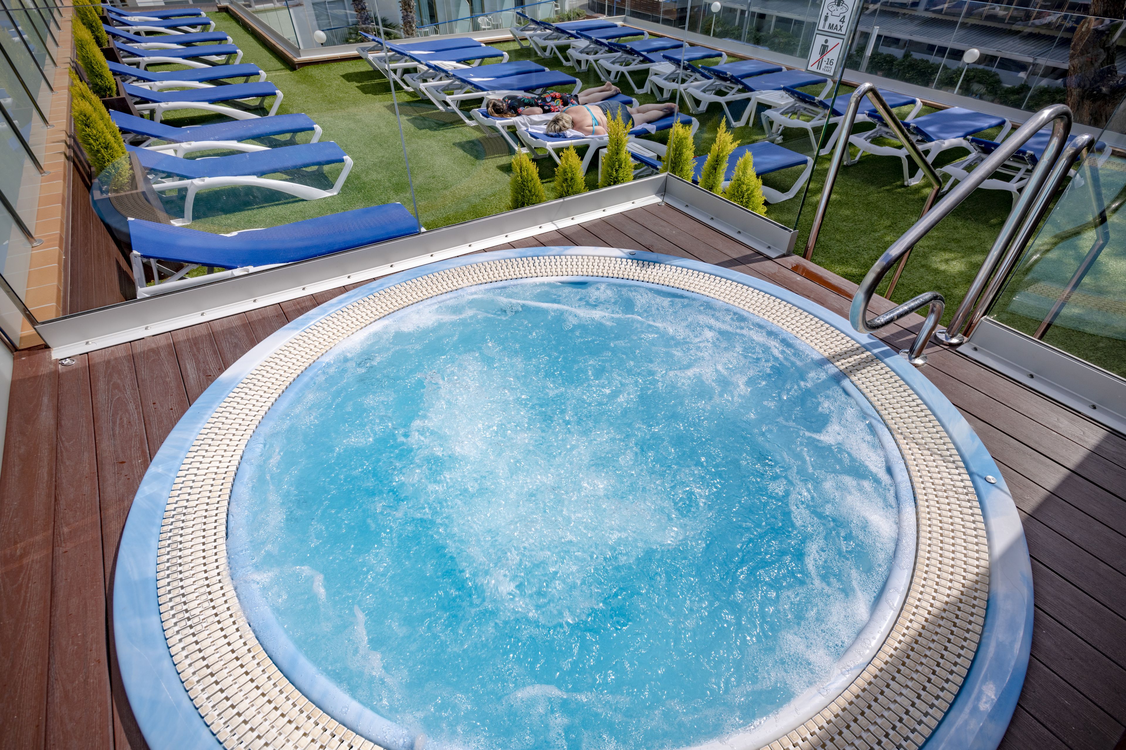 Un jacuzzi rodeado de sillones de descanso en una terraza con barandilla de vidrio, con vista a una zona verde.