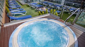 Un jacuzzi rodeado de sillones de descanso en una terraza con barandilla de vidrio, con vista a una zona verde.