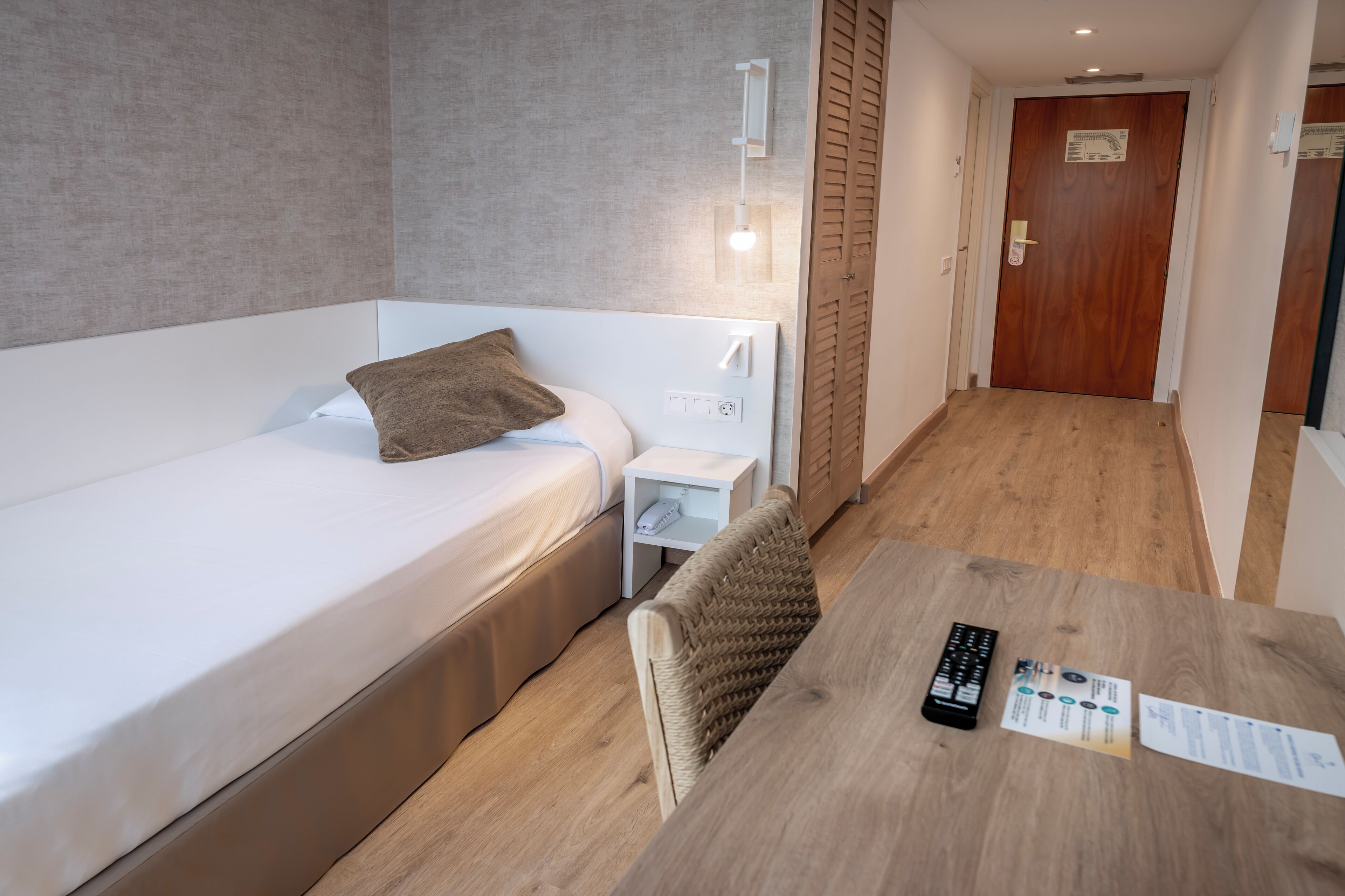 Une chambre d’hôtel avec un lit simple, une table de chevet et une petite table sur laquelle se trouvent une télécommande et une brochure.