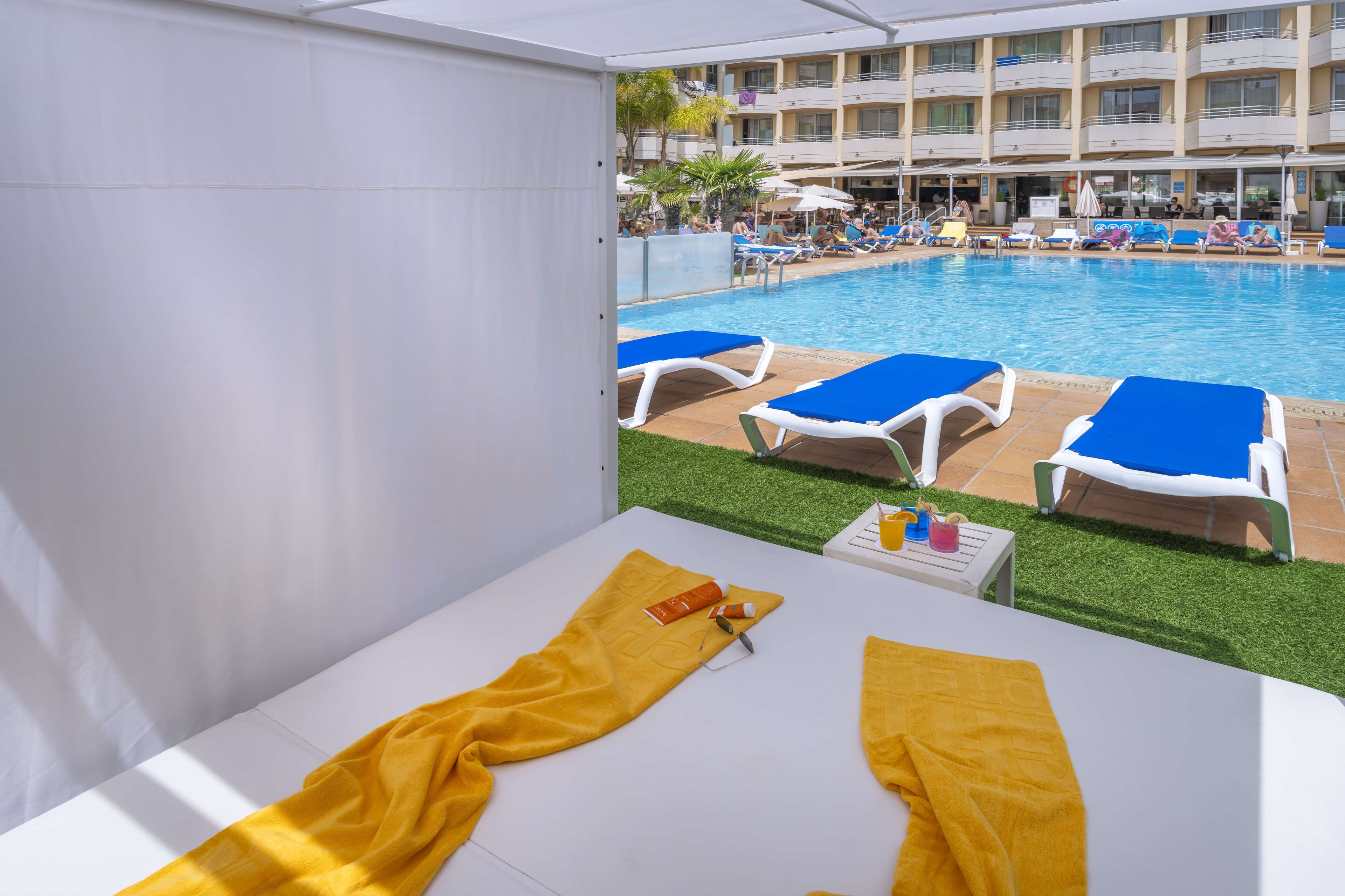 A hotel room with a view of a pool area, including sun loungers and a white table with a yellow towel and toiletries.