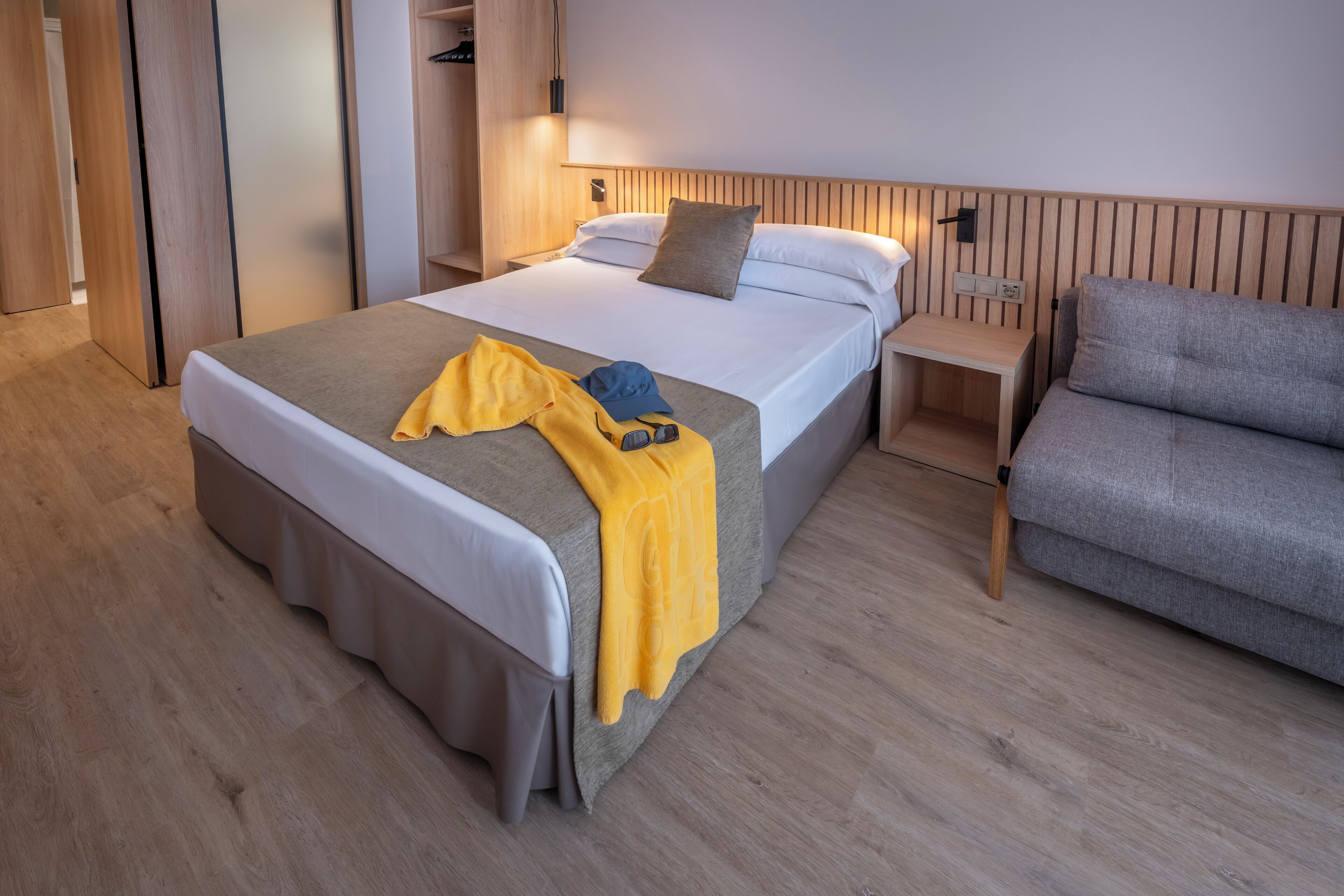 Una habitación de hotel con cama, mesita de noche, sofá gris, suelo de madera y armario.