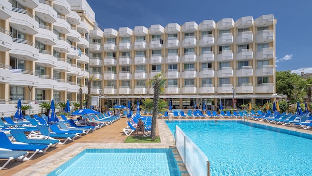 A hotel with a large pool, blue lounge chairs, and a multi-story building with balconies. - Hotel GHT Oasis Tossa & Spa (Tossa de Mar)