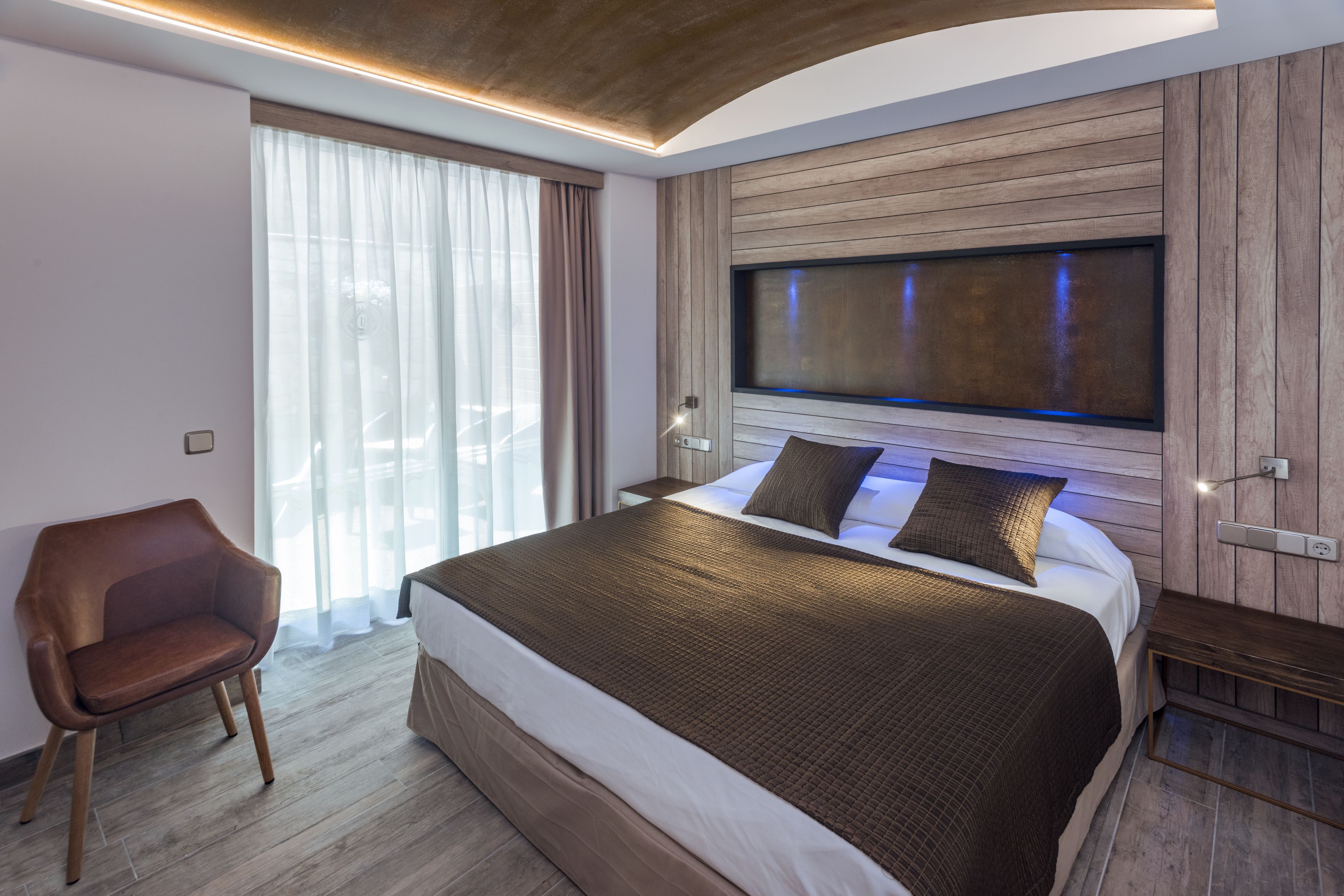 A modern hotel room with a large bed, a wooden headboard, a seating area, and a large window with curtains.