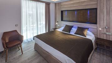 A modern hotel room with a large bed, a wooden headboard, a seating area, and a large window with curtains.