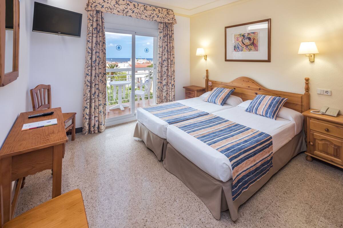 A hotel room with a bed, a desk, a chair, a TV, a balcony with a view, and a painting on the wall.