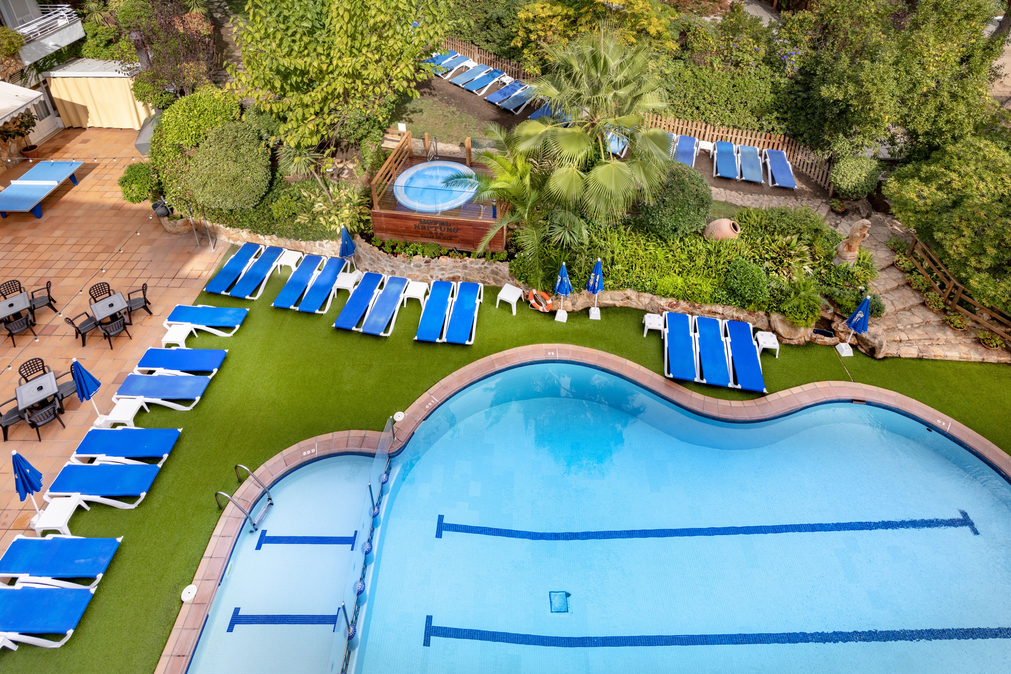 Uma grande piscina com várias espreguiçadeiras e camas de sol dispostas ao redor, cercada por vegetação bem cuidada e uma banheira de hidromassagem.