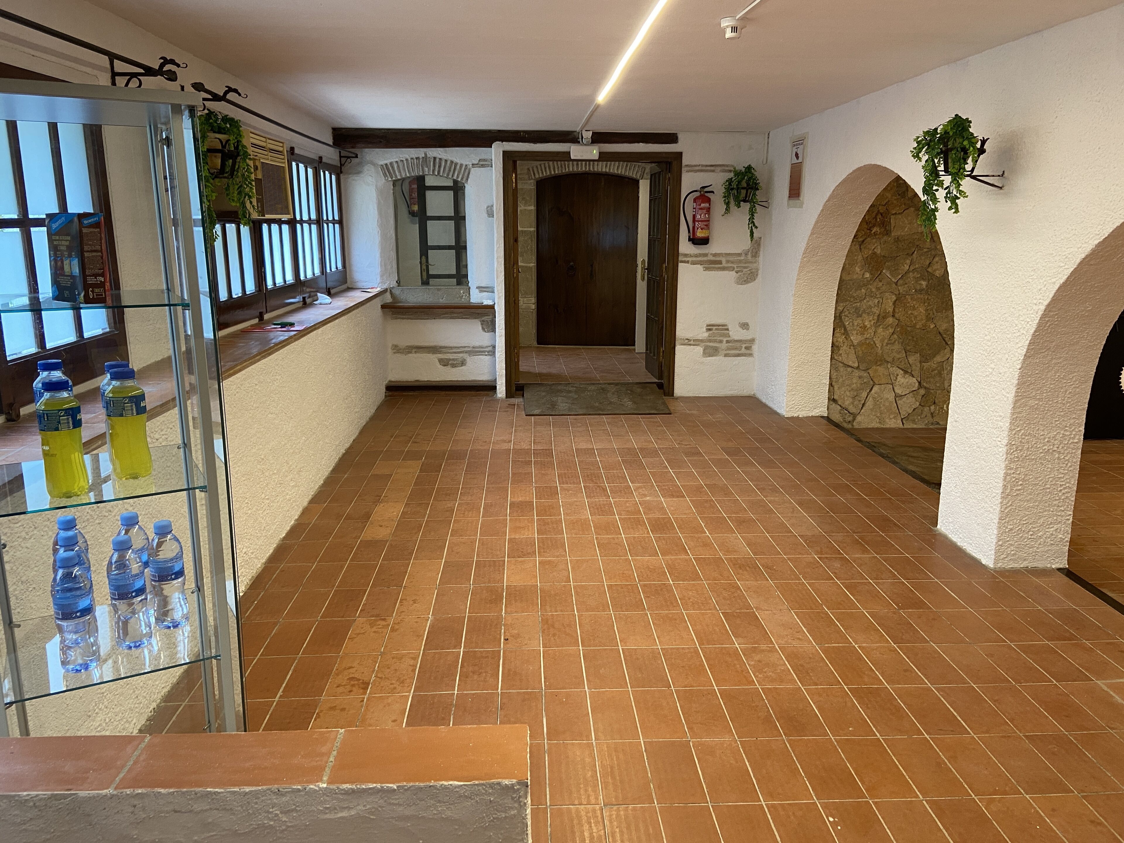 A spacious hallway with tiled flooring, a glass display case with water bottles, a wooden door, and stone arches.