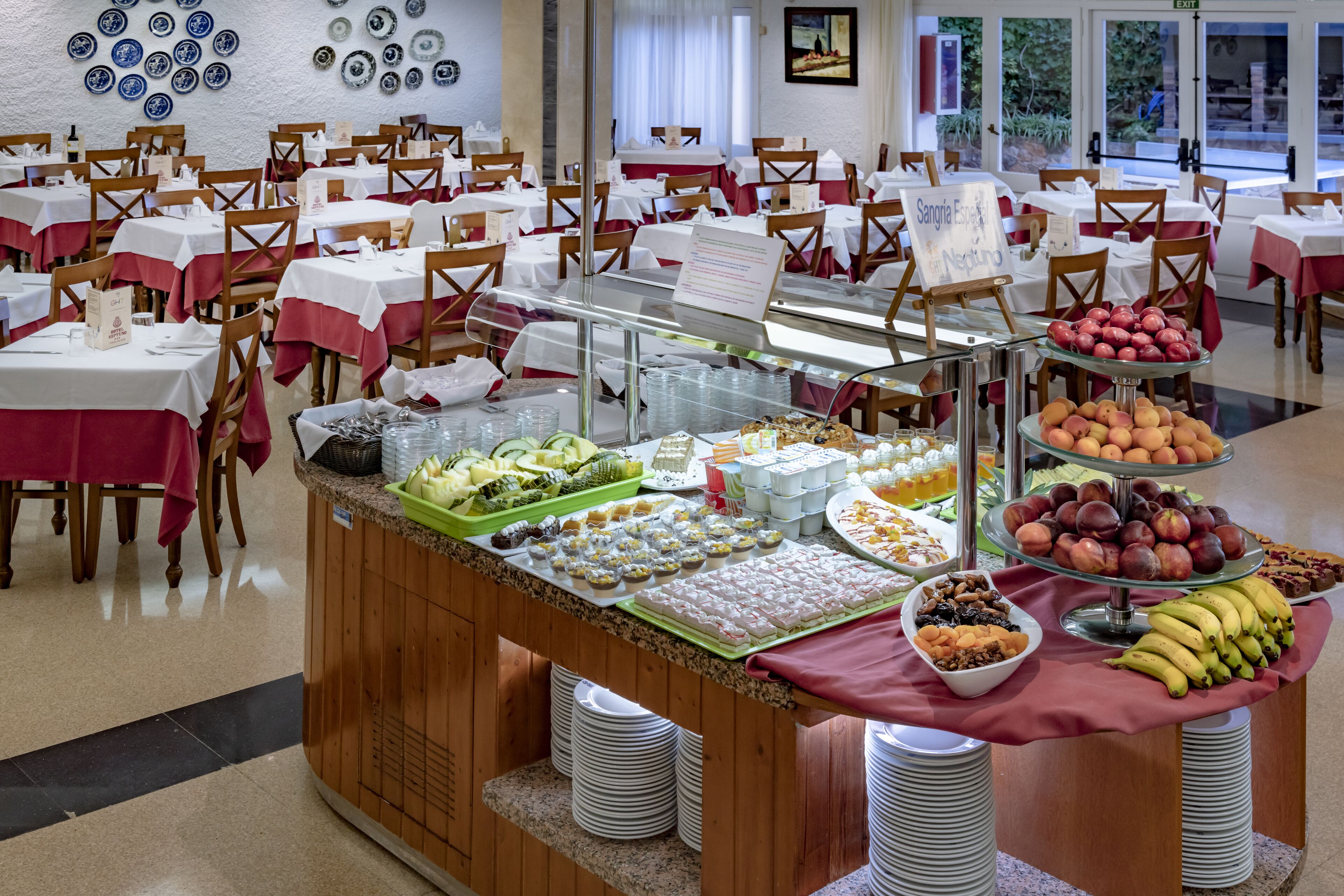 Mesa de buffet con diversas opciones de comida y un cartel que dice 'Sangría & Negativo'. Las mesas están dispuestas con manteles blancos y rojos, y la habitación tiene piso de baldosas y una pared decorada con platos.