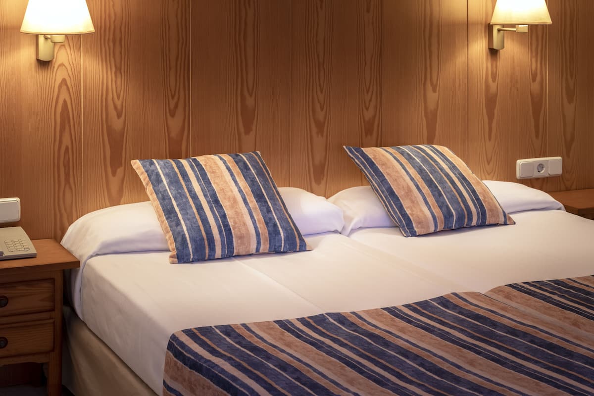 A hotel room with a wooden headboard, two striped pillows, a white bedspread, and a nightstand with a lamp and a phone.