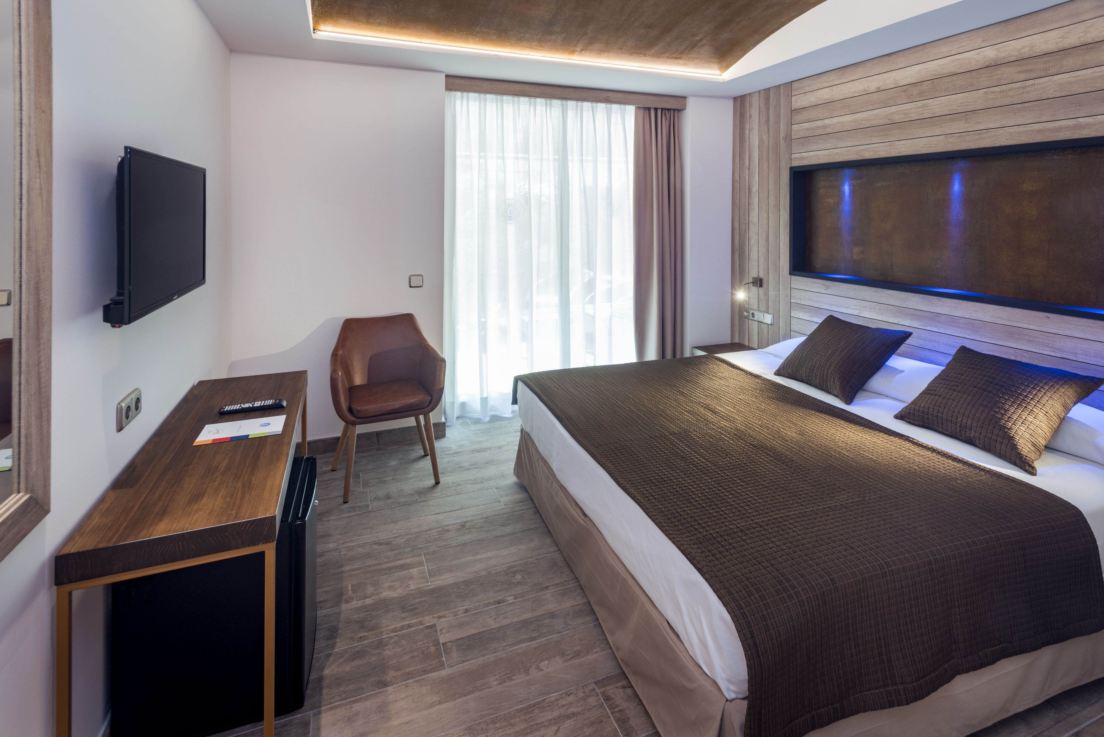 A modern hotel room with a large bed, a desk, a chair, and a TV mounted on the wall.