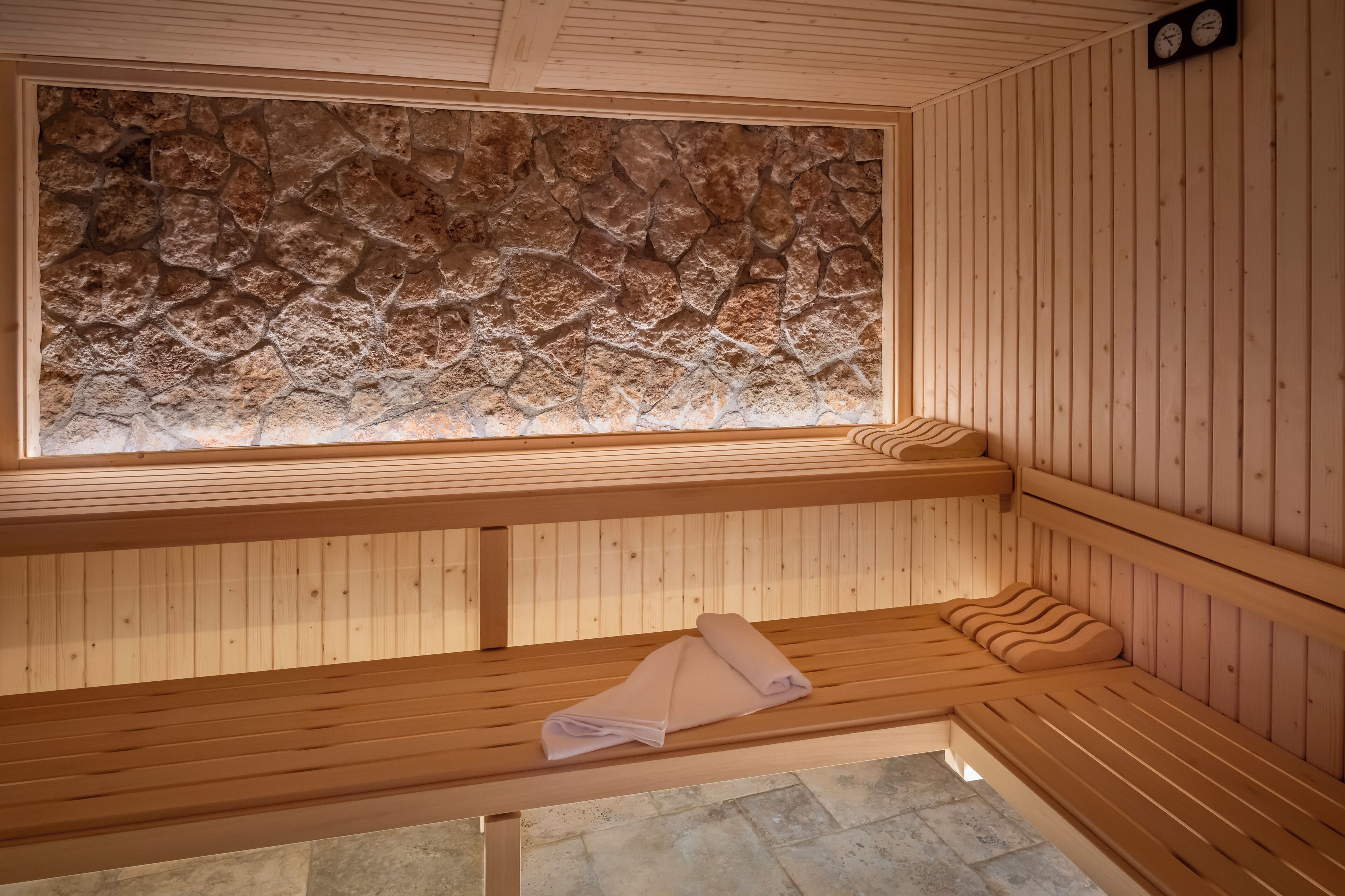 A sauna room with wooden benches, a stone wall, and a clock on the wall.