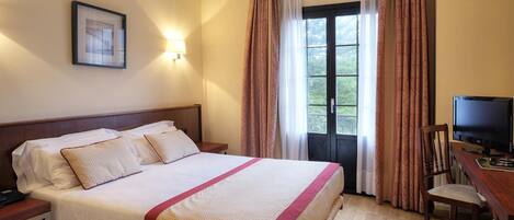 A hotel room with a bed, a desk with a television, and a window with curtains.