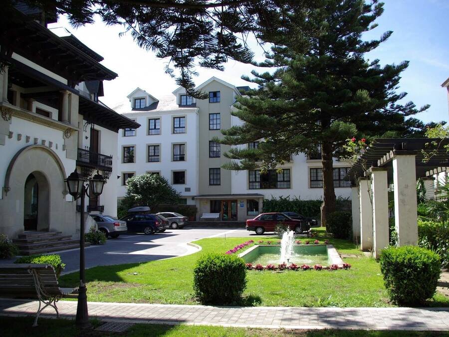 Un patio con una fuente, coches aparcados y un edificio con varias ventanas y balcones.