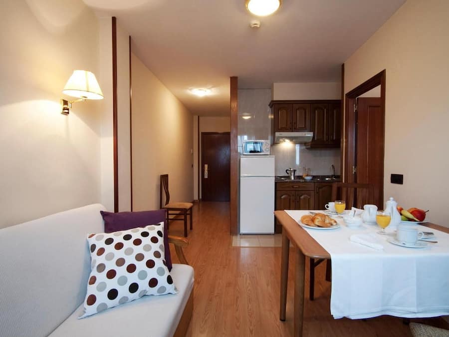 Una habitación acogedora con sofá, una mesa de comedor lista para el desayuno, una cocina al fondo y una lámpara en la pared.