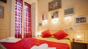 Habitación de hotel con una cama grande, cortinas rojas y blancas, luces fijadas a la pared y cuadros enmarcados.