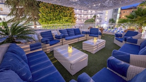 A rooftop lounge area with blue upholstered sofas and wicker coffee tables, surrounded by greenery and a white railing. - GHT Balmes, Hotel, Aparthotel & SPLASH (Calella)
