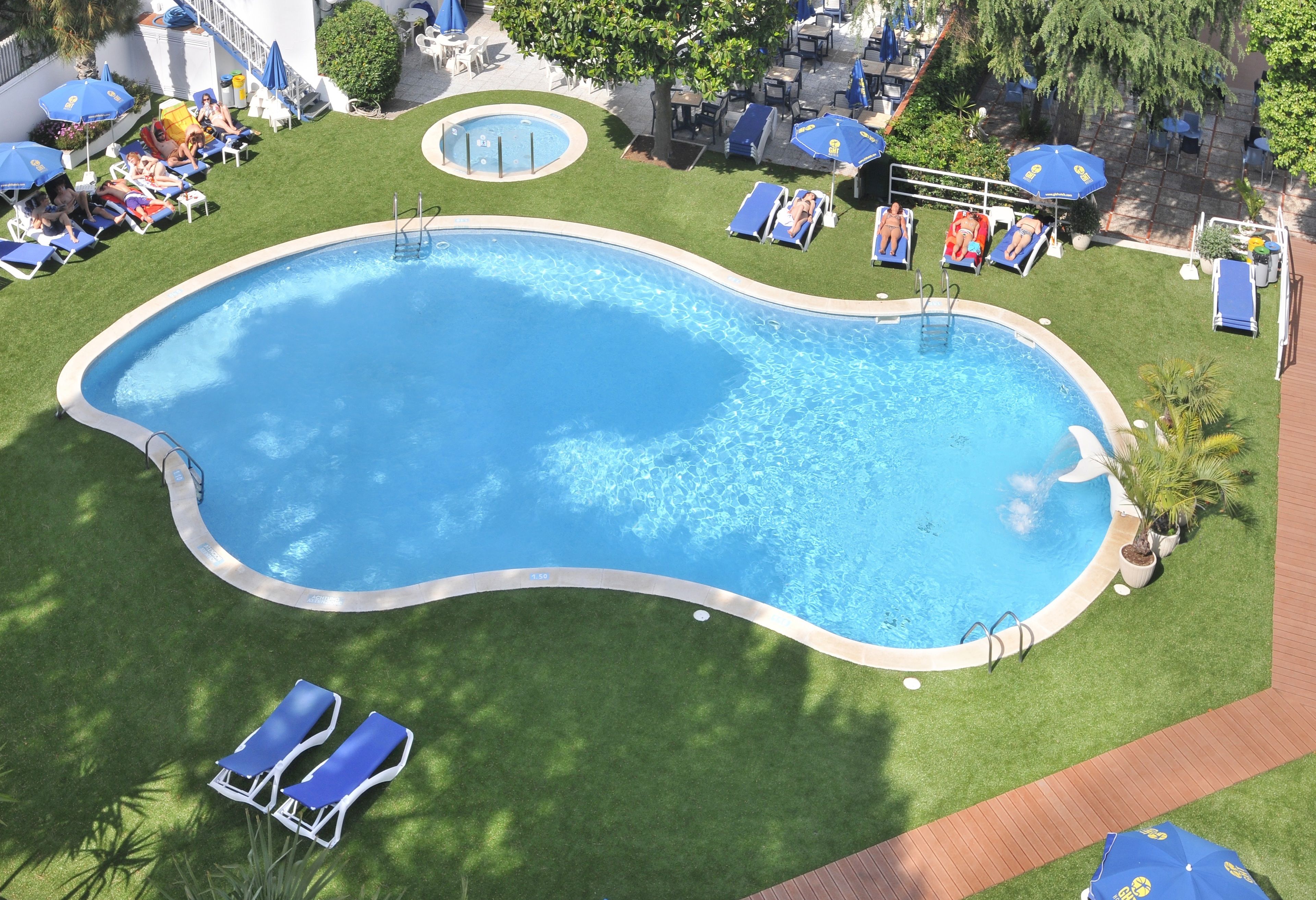 An aerial view of a swimming pool with multiple sun loungers and umbrellas, surrounded by well-maintained grass and trees.