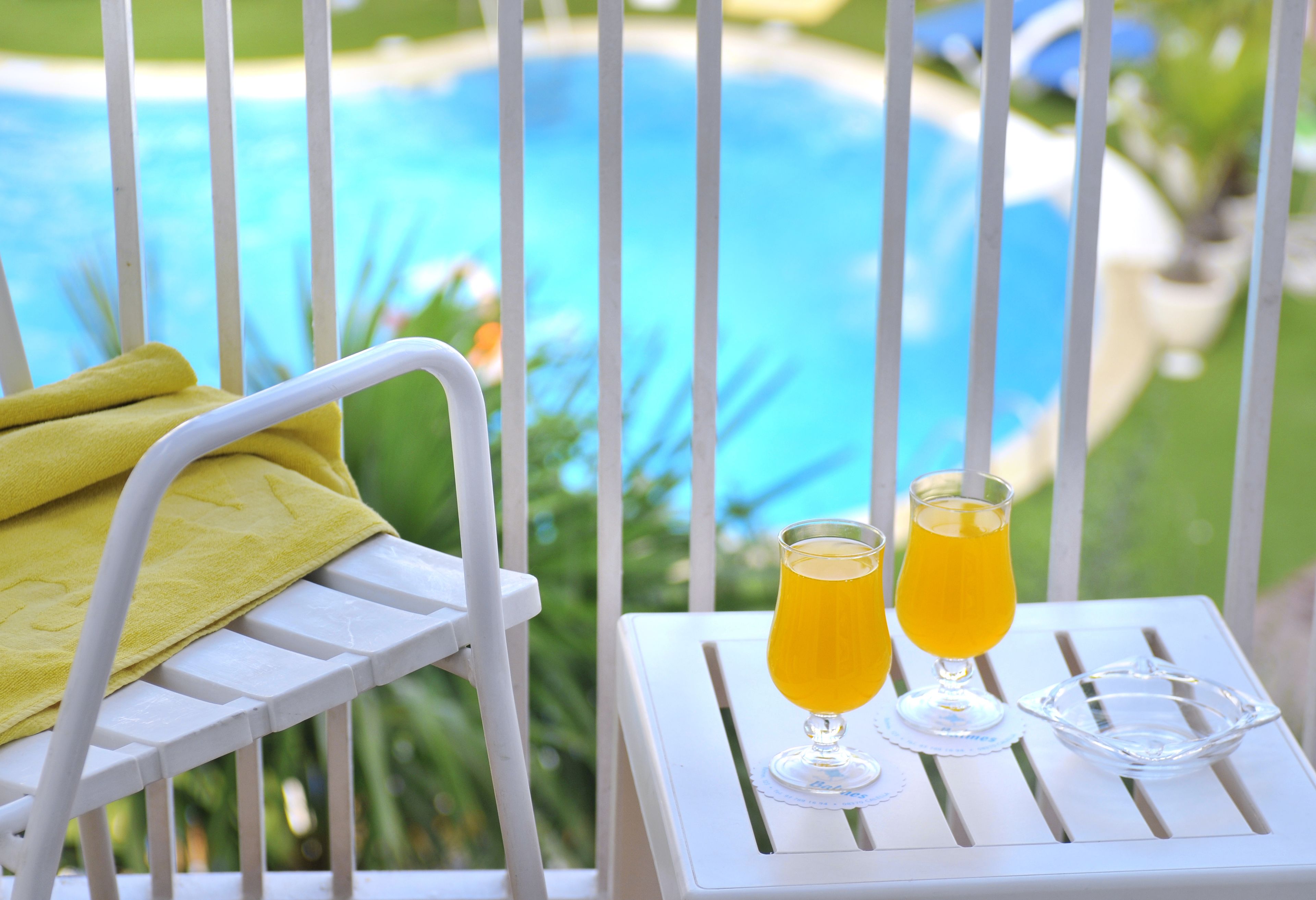 Una silla de metal blanca con una toalla amarilla, dos vasos de zumo de naranja y un bol de vidrio transparente sobre una mesa blanca con vista a la piscina.