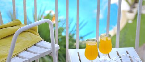 Una silla de metal blanca con una toalla amarilla, dos vasos de zumo de naranja y un bol de vidrio transparente sobre una mesa blanca con vista a la piscina.