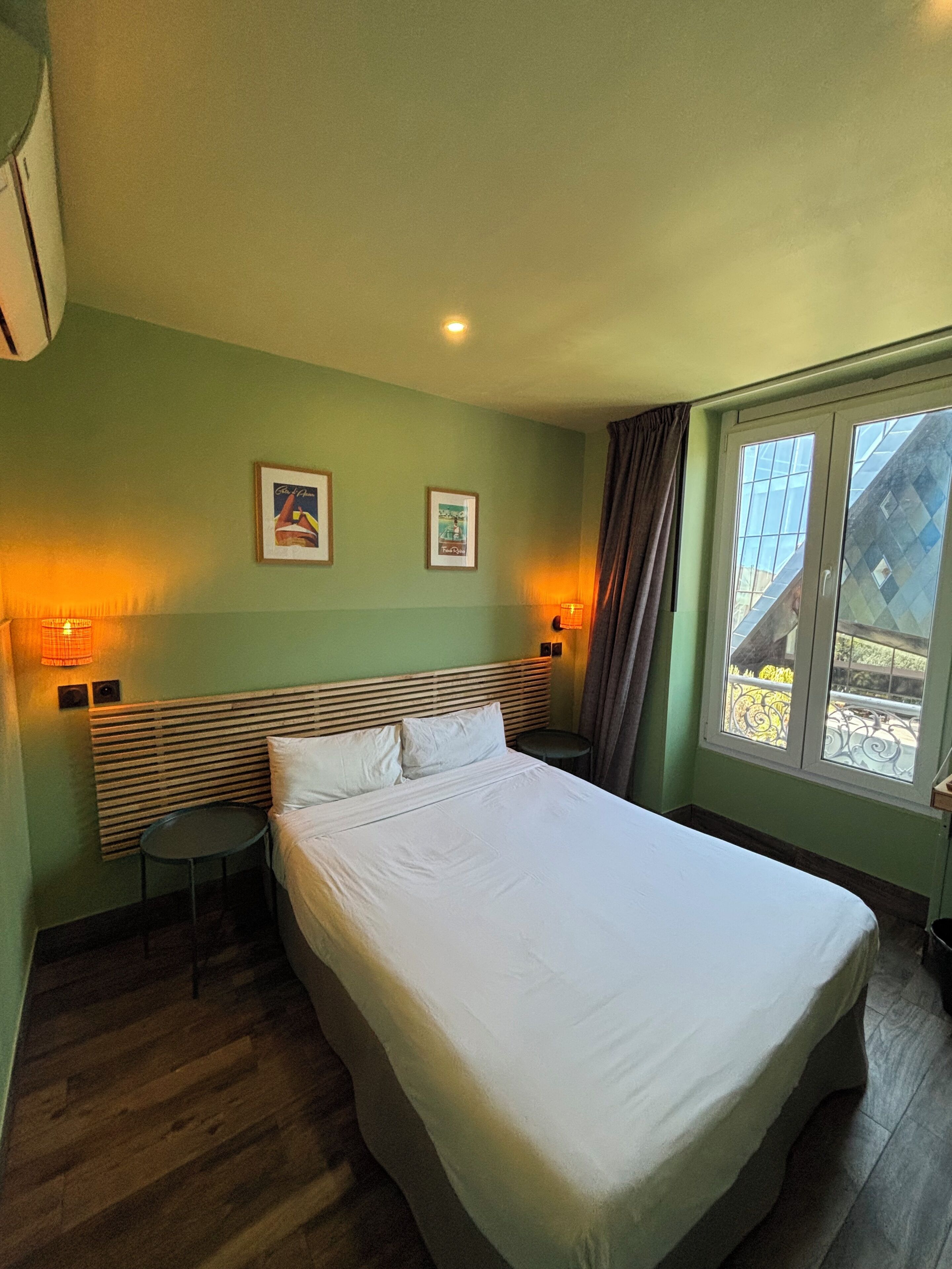 Une chambre d’hôtel avec un lit, deux lampes fixées au mur, une petite table ronde et une fenêtre donnant sur des immeubles.