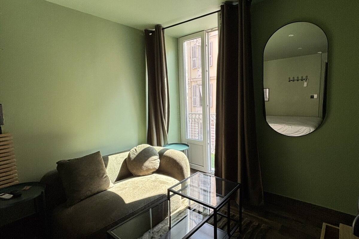 Una habitación con sofá, una mesa de centro de vidrio, un espejo y vistas a edificios a través de una ventana.
