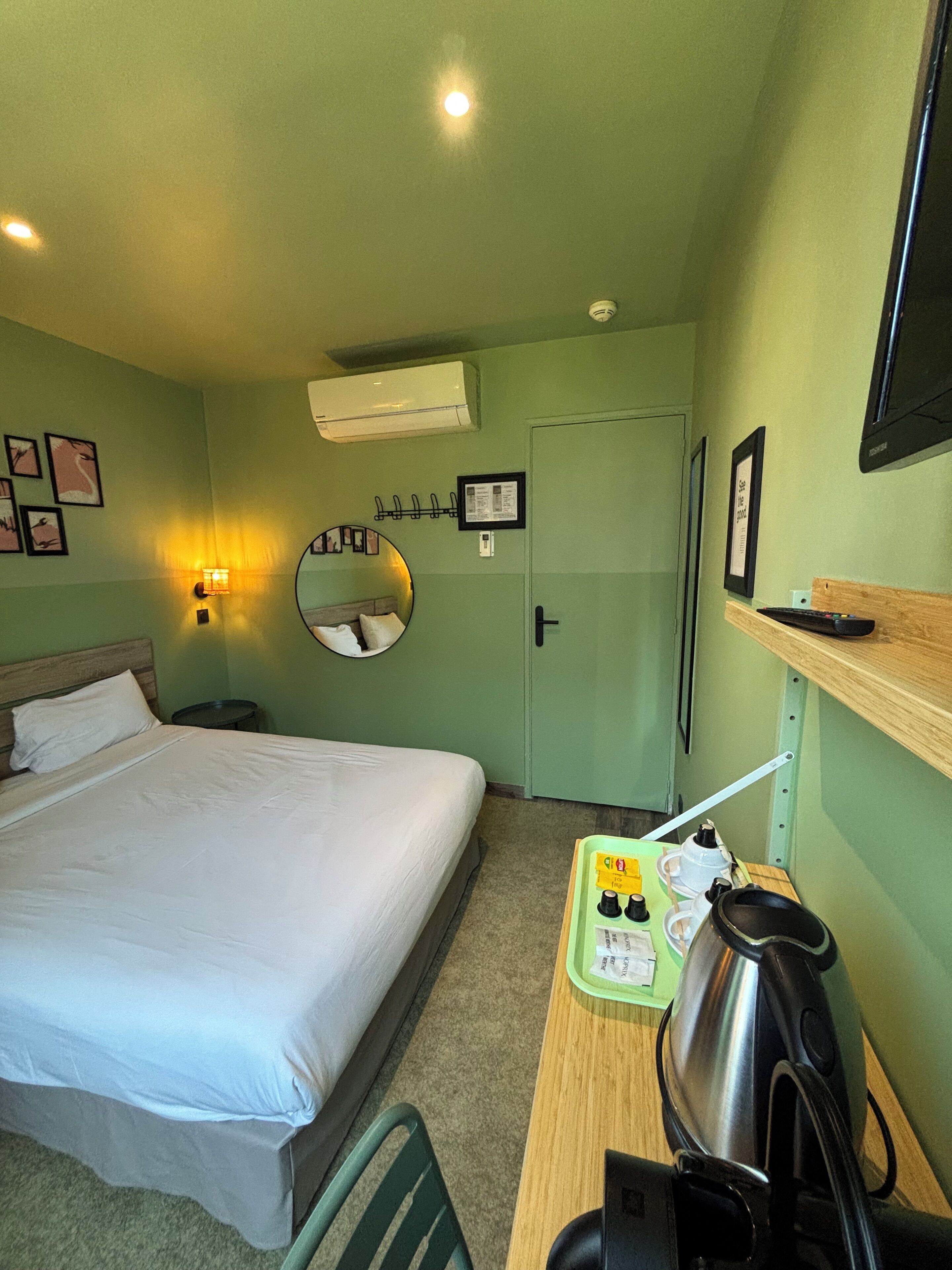 A hotel room with a bed, a desk with a kettle, a mirror, and a TV on the wall.