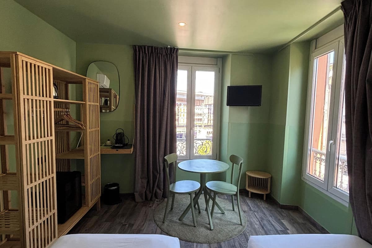 Una habitación con dos camas, un armario de madera, una mesa redonda con sillas, un taburete pequeño, un televisor y una ventana con cortinas.