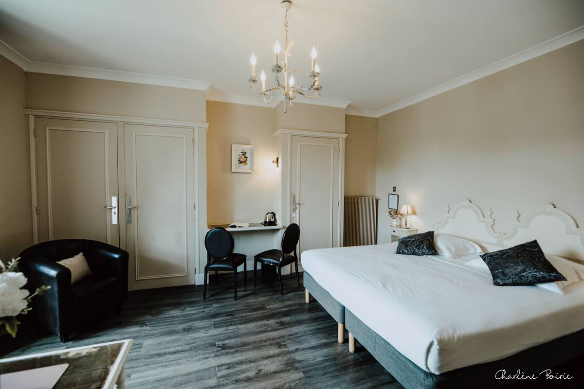 Habitación de hotel con una cama grande, un escritorio, dos sillas, un sillón de cuero negro, una araña y un radiador.