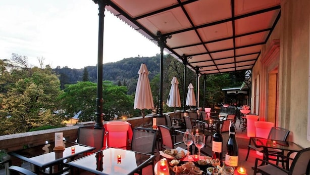 Un área de comedor al aire libre con mesas y sillas, una mesa iluminada en rojo con comida y bebidas, y un paisaje de árboles y colinas al fondo.