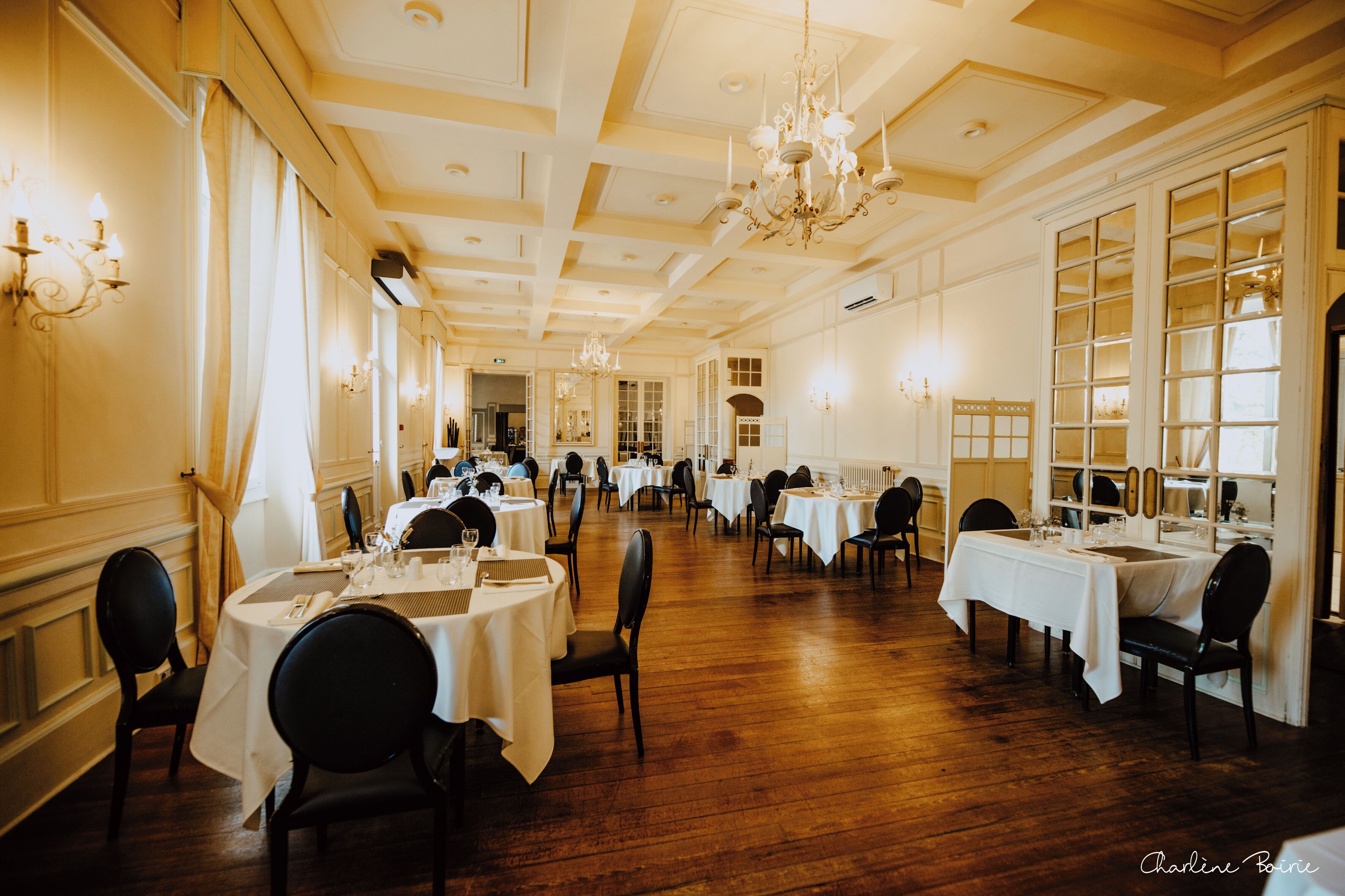 A spacious dining hall with multiple round tables set for dining, elegant chandeliers, and large windows allowing natural light.