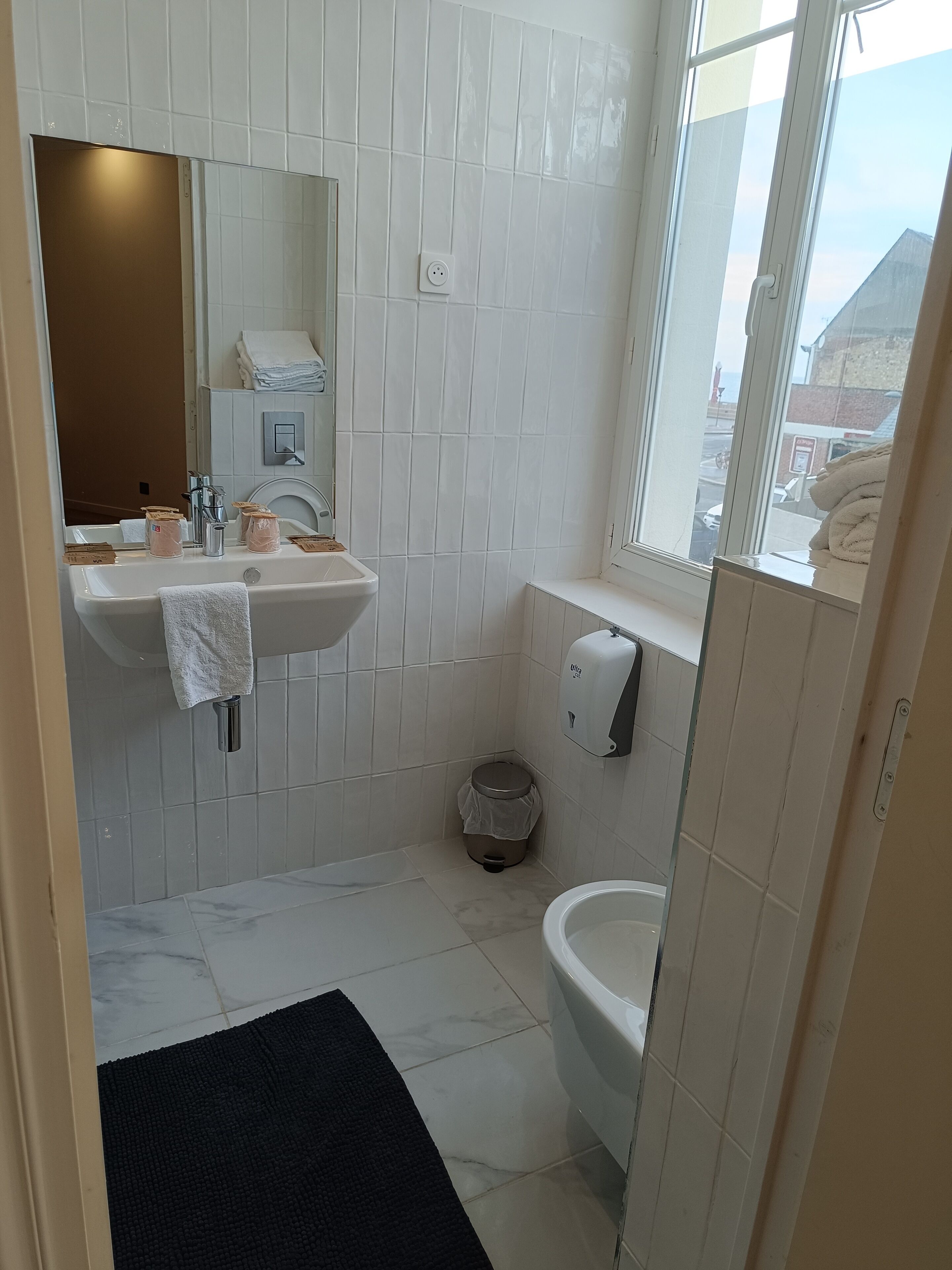Een badkamer met een wit wastafelmeubel, spiegel en toilet, een zwarte badmat en een raam met uitzicht op de omgeving.