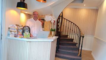 Un espace d’accueil avec un comptoir, un ordinateur portable et une orchidée en pot. On y trouve un escalier avec une rampe en bois et un parquet disposé en motif à chevrons.