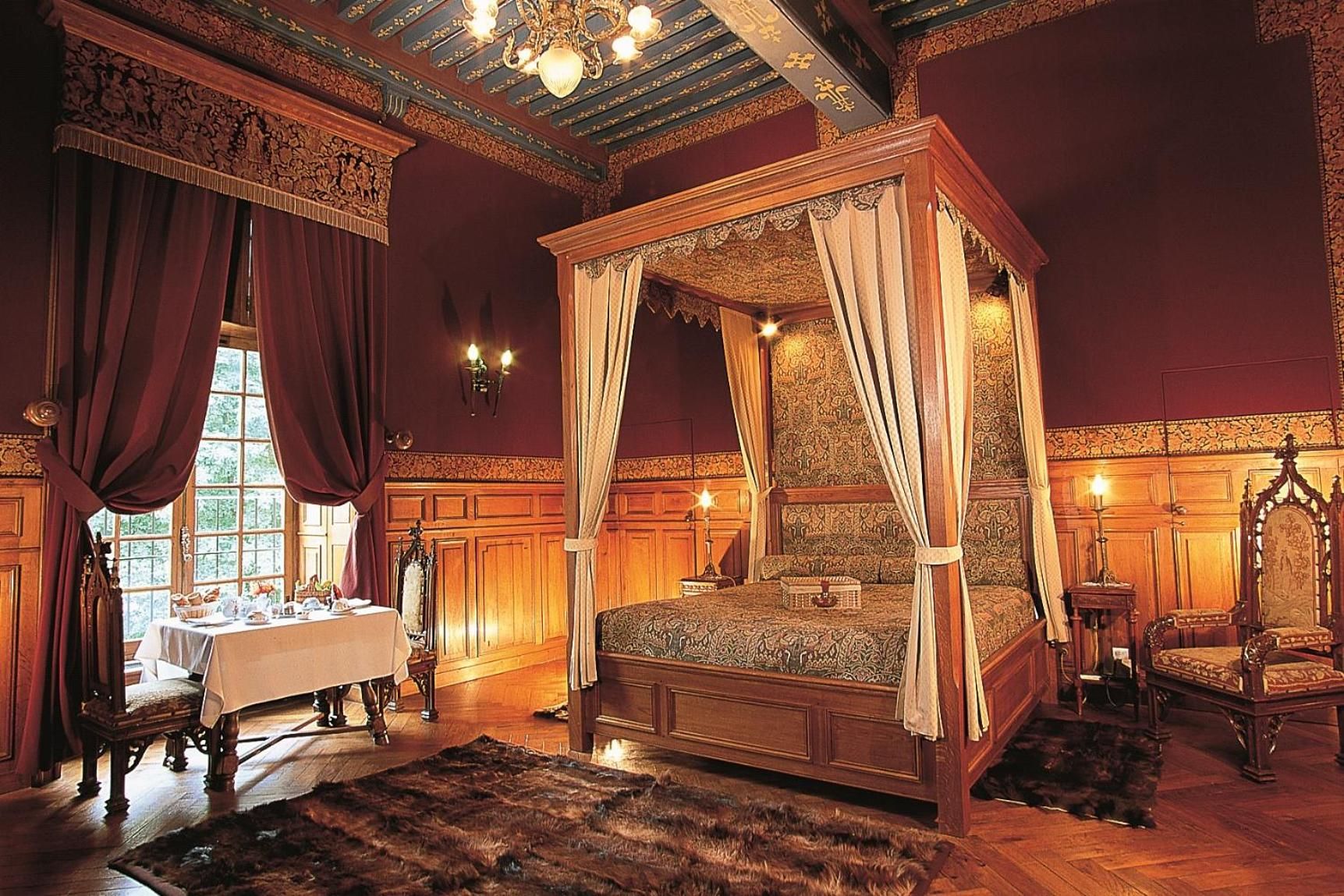 A four-poster canopy bed with a wooden frame and draped curtains, a table with a white tablecloth, and a chair with a dark cushion.