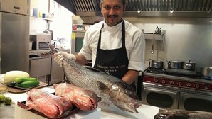 Un chef se tient dans une cuisine, tenant un gros poisson. On peut voir divers ustensiles de cuisine et ingrédients autour de lui.