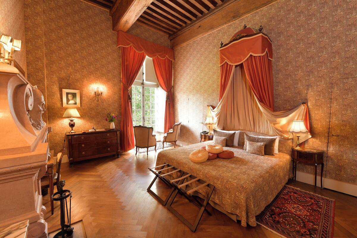 Ein Schlafzimmer mit einem großen Bett, Holzboden, einem Schminktisch, einem Sessel und einem Fenster mit Vorhängen.