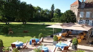 Un espace extérieur aménagé en terrasse, avec des tables et des chaises, entouré de verdure et avec un bâtiment en arrière-plan.