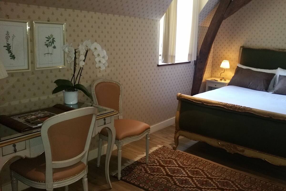 Ein Schlafzimmer mit einem Bett, einem Schreibtisch mit Stuhl, einem Nachttisch, einer Lampe und einem Fenster mit Vorhängen.