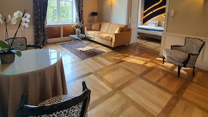 Un salon comprenant un canapé, des fauteuils, une table basse en verre et une table à manger. Il y a une fenêtre avec des rideaux, une lampe et un tableau au mur.