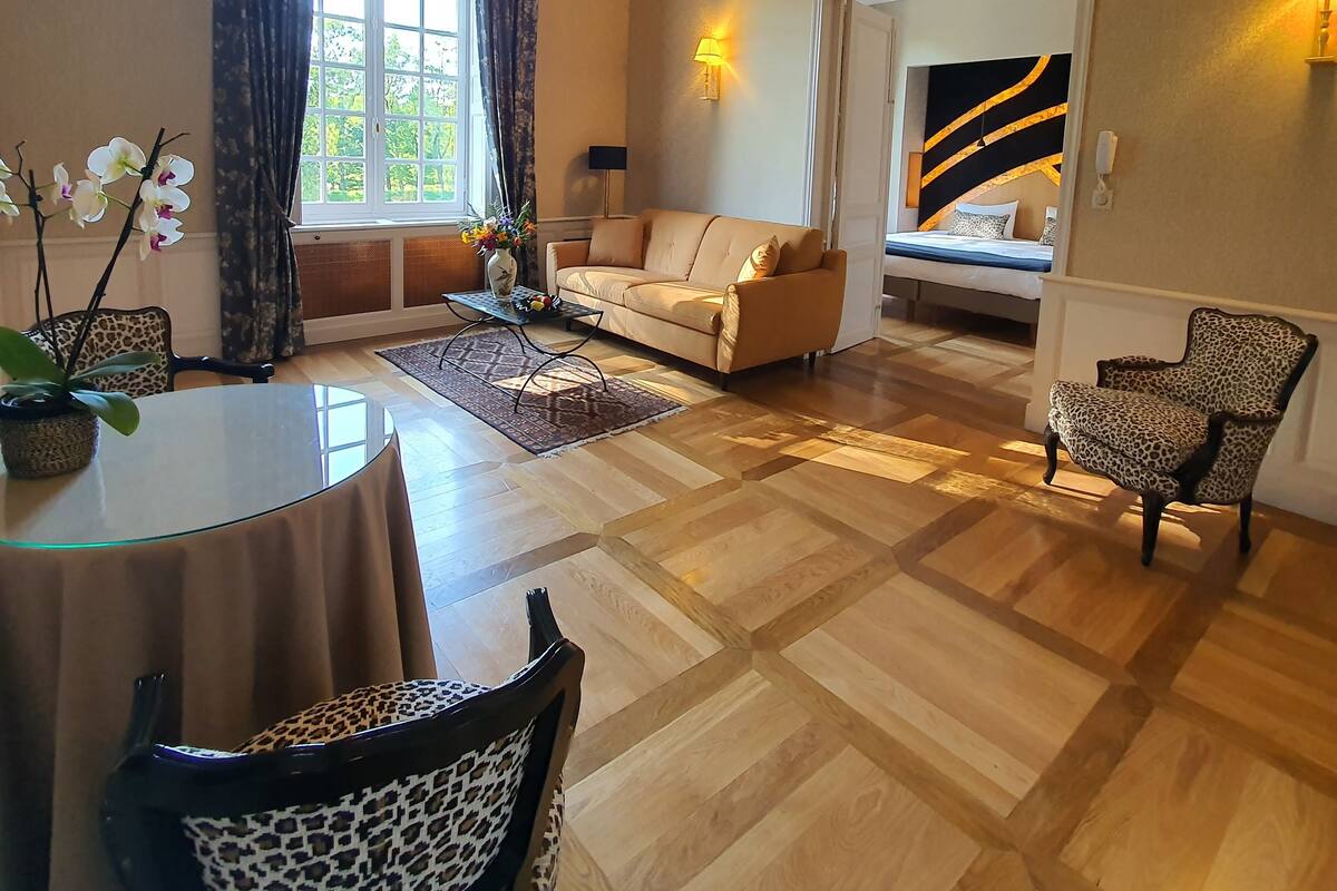 Ein Wohnzimmer mit einer Couch, Sesseln, einem Glastisch und einem Esstisch. Es gibt ein Fenster mit Vorhängen, eine Lampe und ein Gemälde an der Wand.