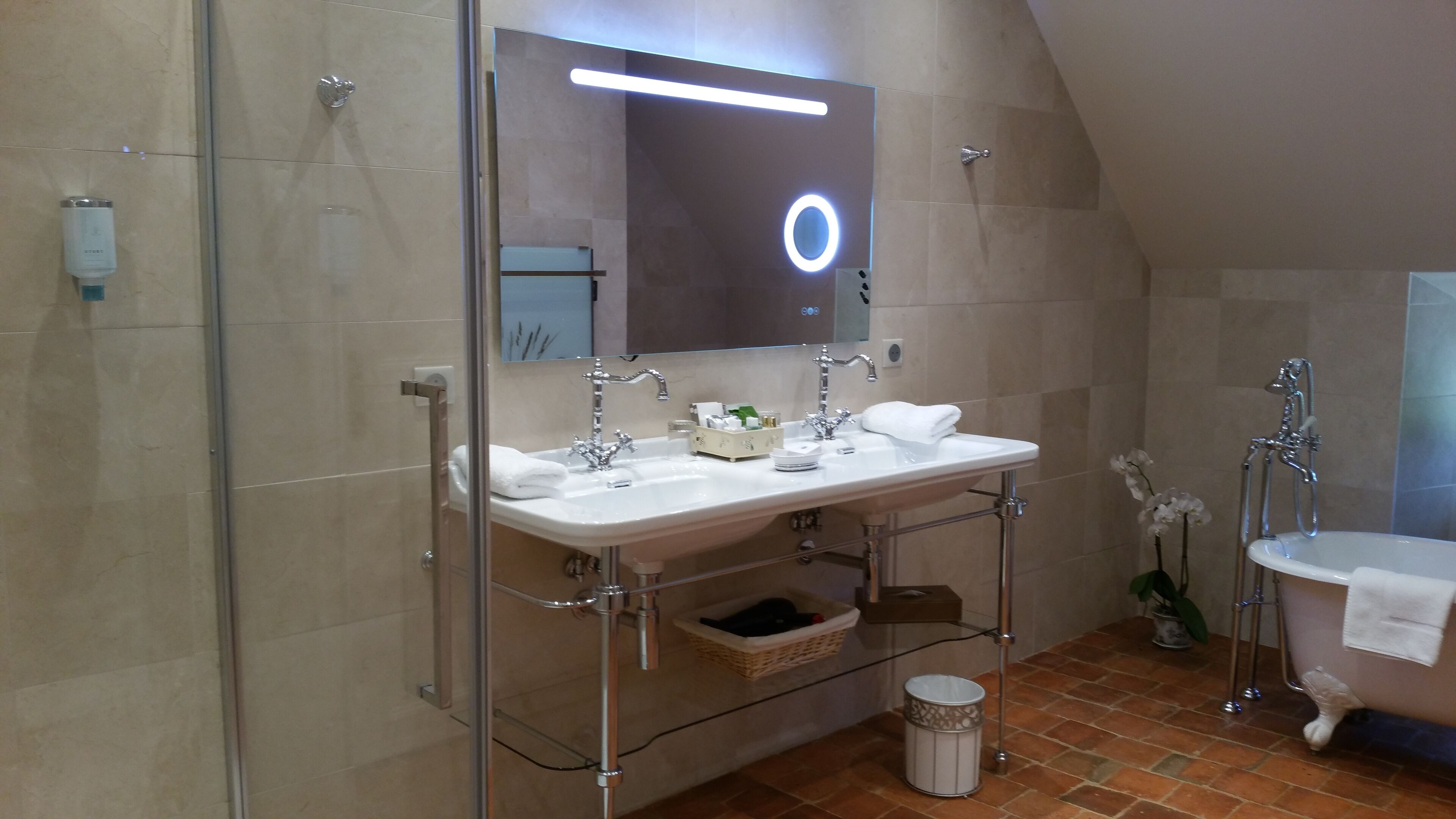 Un baño moderno con cabina de ducha de vidrio, tocador doble lavamanos, bañera independiente y espejo con luz circular.