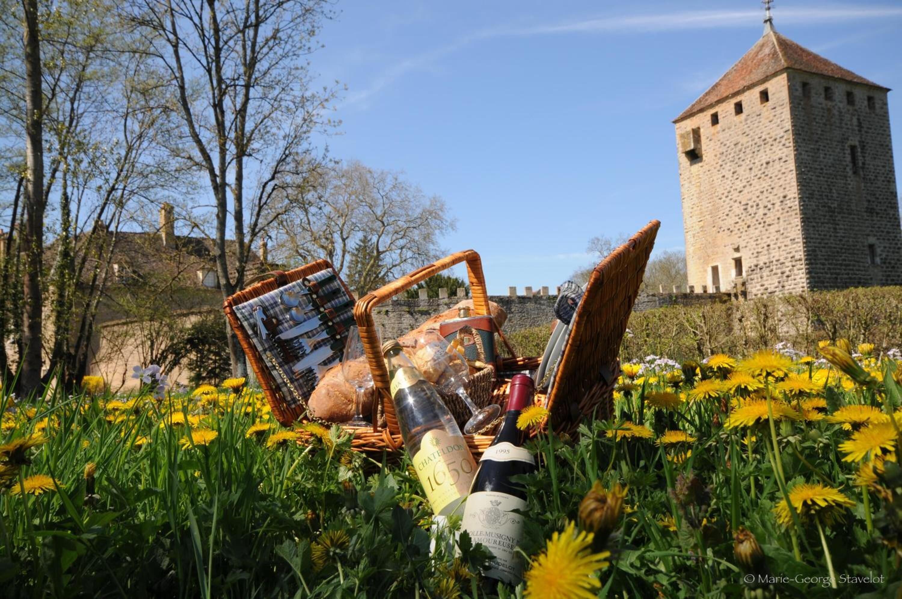 Ein Picknick mit Korb, Flasche und Brot in einem Feld voller Löwenzahn, im Hintergrund ein Steinturm.