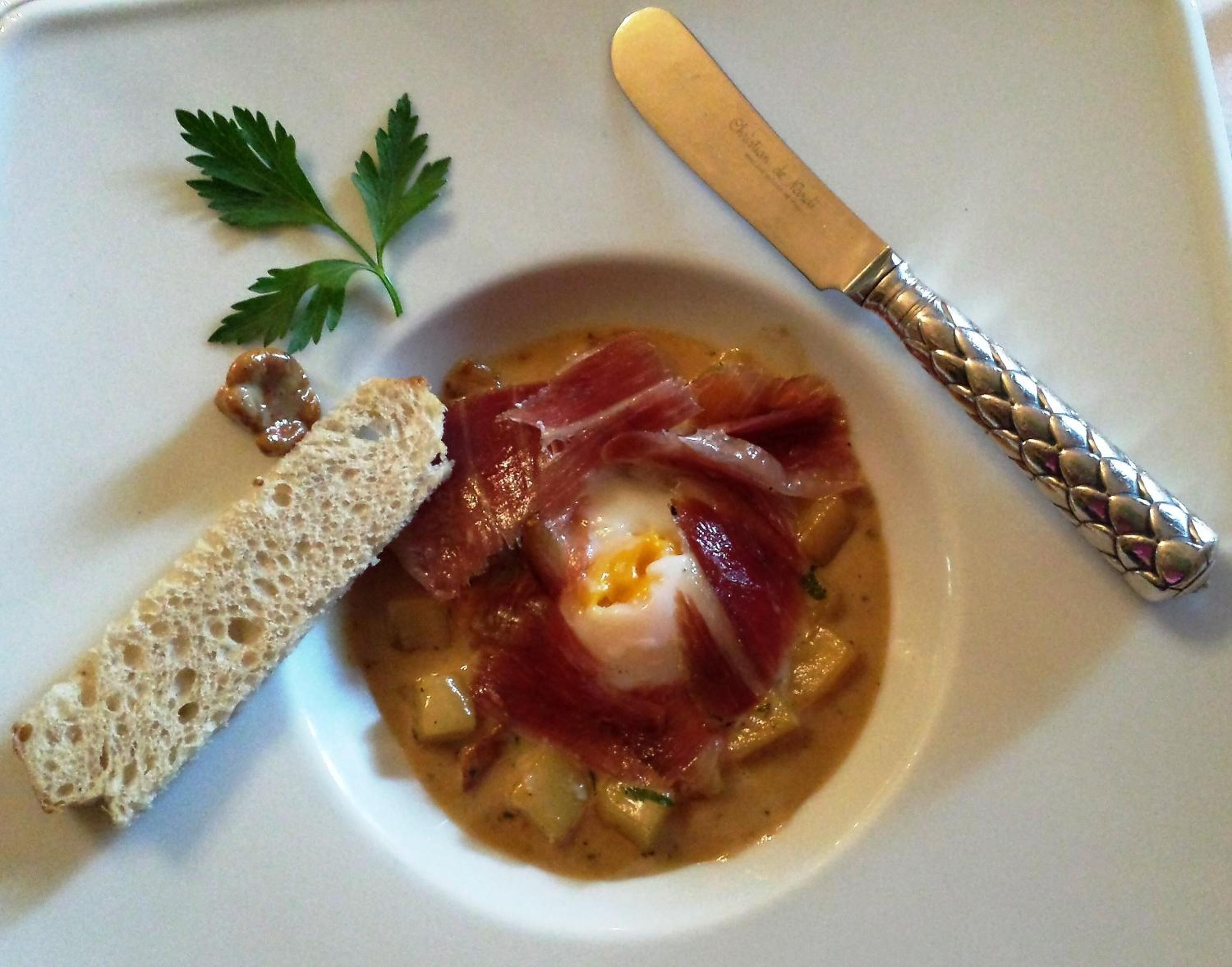 A bowl of soup with a poached egg, prosciutto, and bread on a white plate.