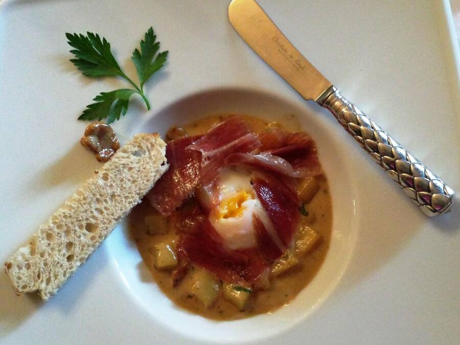 A bowl of soup with a poached egg, prosciutto, and bread on a white plate.