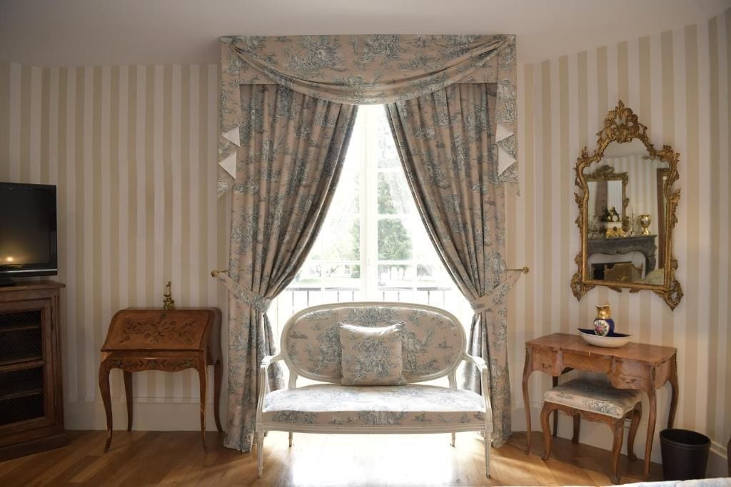 A room with a large window, a television on a wooden cabinet, a sofa, a small table, and a mirror on the wall.