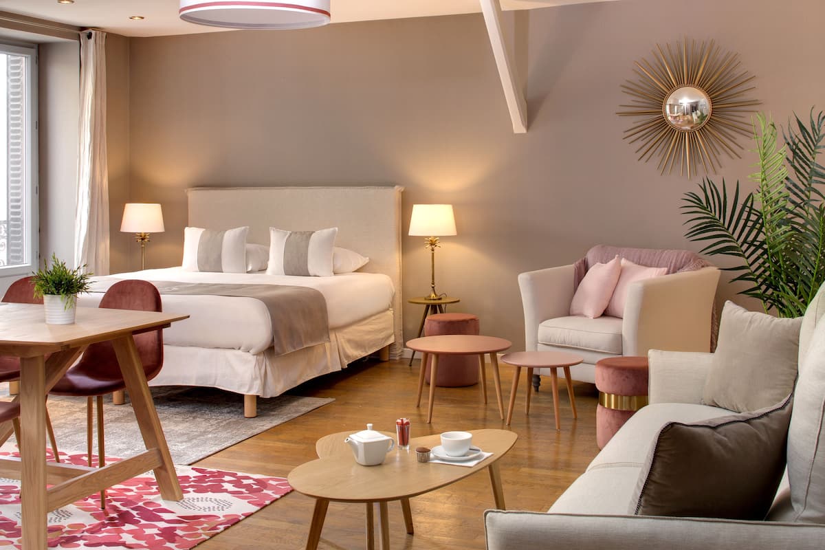 A hotel room with a bed, two chairs, a sofa, a small table, and a coffee table. There is a lamp on a side table and a plant in the corner.