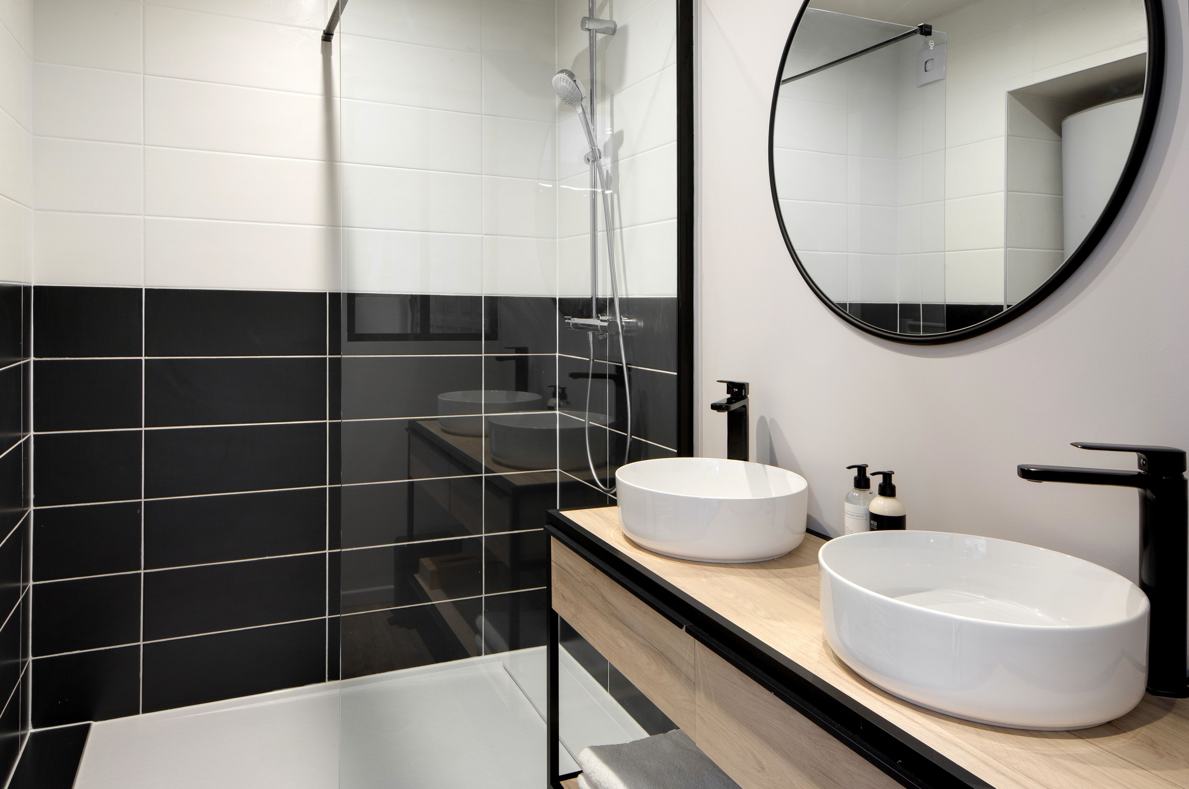 A modern bathroom with black and white tiles, a round mirror, two sinks, and a shower.